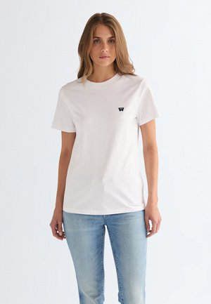 Vrouw met lichtbruin haar, gekleed in een eenvoudige witte korte mouw T-shirt en lichtblauwe spijkerbroek, staand tegen een effen witte achtergrond.