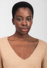 Beige, zacht gebreid sweater met een diepe v-hals en een gestructureerde afwerking. Model heeft kort haar en subtiele oorbellen.