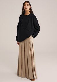 Juodas per didelis megztinis su giliu V formos iškirpimu, derinamas su ilgu smėlinės spalvos plisuotu sijonu. Apranga pasižymi tekstūruota medžiaga ir subtiliu drapavimu.