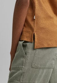 T-shirt marron à manches courtes avec fentes sur les côtés, associé à un pantalon vert olive avec des poches à rabat et un tissu texturé.