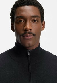 Pull noir à col zippé en tissu texturé, avec un col côtelé et une fermeture éclair métallique, porté par un homme aux cheveux courts et bouclés.