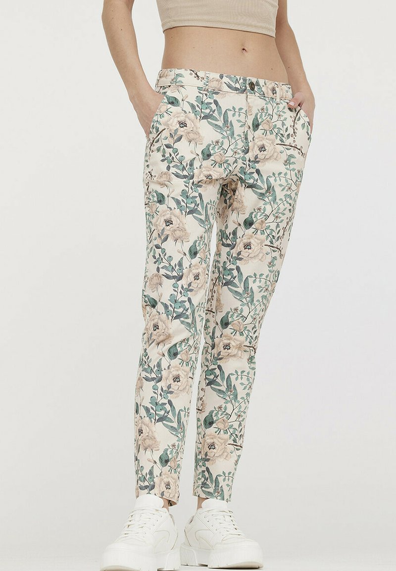 Pantalons à imprimé floral en crème avec des feuilles vertes et des fleurs beiges. Présente deux poches latérales et une coupe ajustée. Portés avec des baskets blanches.