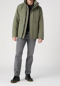 Homme portant une veste olive, un pull noir, un jean gris et des bottes en cuir noir se tenant devant un fond blanc uni.
