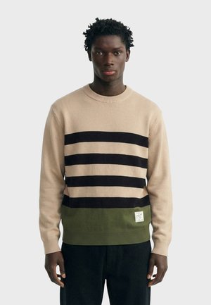 Mann trägt einen beigen Pullover mit drei schwarzen horizontalen Streifen und grünem Saum, steht vor einem einfarbigen hellen Hintergrund.