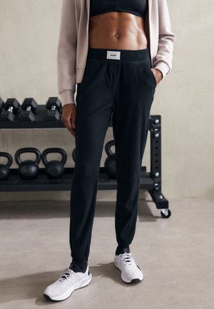 Sportowiec ubrany w czarne prążkowane spodnie dresowe, czarny stanik sportowy, beżową kurtkę i białe sneakersy stoi w pobliżu kettlebelli i hantli na siłowni.