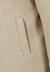 Smėlio spalvos vilnos mišinio audinys su minkšta tekstūra, kuriame yra stačiakampio formos kišenė, įkomponuota į siūlę, siekiant sukurti tvarkingą išvaizdą.