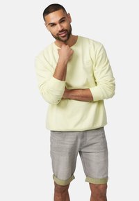 Felpa gialla chiaro con scollatura rotonda, maniche lunghe e logo ricamato. Abbinata a pantaloni corti grigio chiaro arrotolati, che mostrano un look casual.