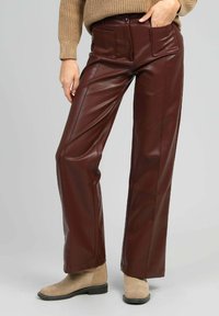 Personne portant un pantalon en cuir marron foncé, des bottines beige à la cheville et un pull en tricot beige, avec une main dans la poche et l'autre détendue.