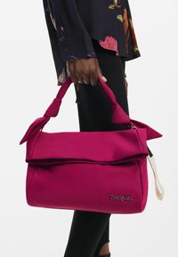 Tasche aus weichem, pinkfarbenem Stoff mit umschlagbarer Klappe, geknoteten Griffen und geprägt "Desigual"-Logo an der Seite.
