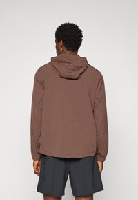 Mand med kort, krøllet hår iført en brun hættetrøje og mørkegrå shorts, der står overfor en ensartet lys baggrund.
