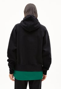 Felpa nera con una vestibilità comoda, dotata di cappuccio con lacci. Sovrapposta a una maglietta verde, con un tessuto testurizzato e polsini a coste.