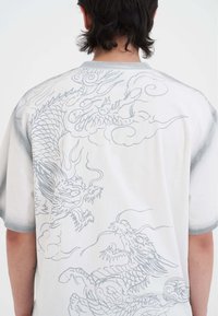Krátký rukáv košile z bílé látky s šedými vzory draka a mraků na zádech, kulatým výstřihem a jemnými šedými akcenty.