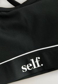 Čierny športový top vyrobený z hladkej, elastickej tkaniny. Má biely akcentový pruh a slovo "self." vytlačené bielou farbou na páske.