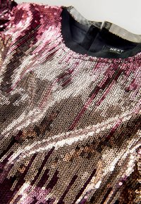 Top zdobený flitry s gradientem růžových a zlatých tónů, s černým výstřihem a třpytivou texturou po celém povrchu. Žádné viditelné vzory ani kovové prvky.