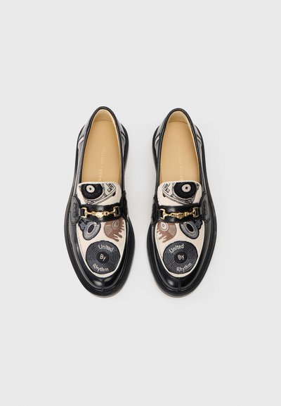 Ett par svarta läderloafers med beige broderade kanvastoppar som pryder skivspelare och texten "United By Rhythm", i detalj med guldfärgad metall.