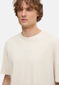 Camiseta de algodón beige con cuello redondo, mangas cortas y un sutil logo grabado en el pecho. Textura suave, corte relajado.