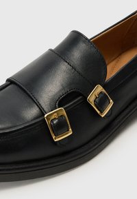 Mocassini in pelle nera con doppia fibbia in hardware color oro, superficie liscia e design a punta rotonda con suola a profilo basso.