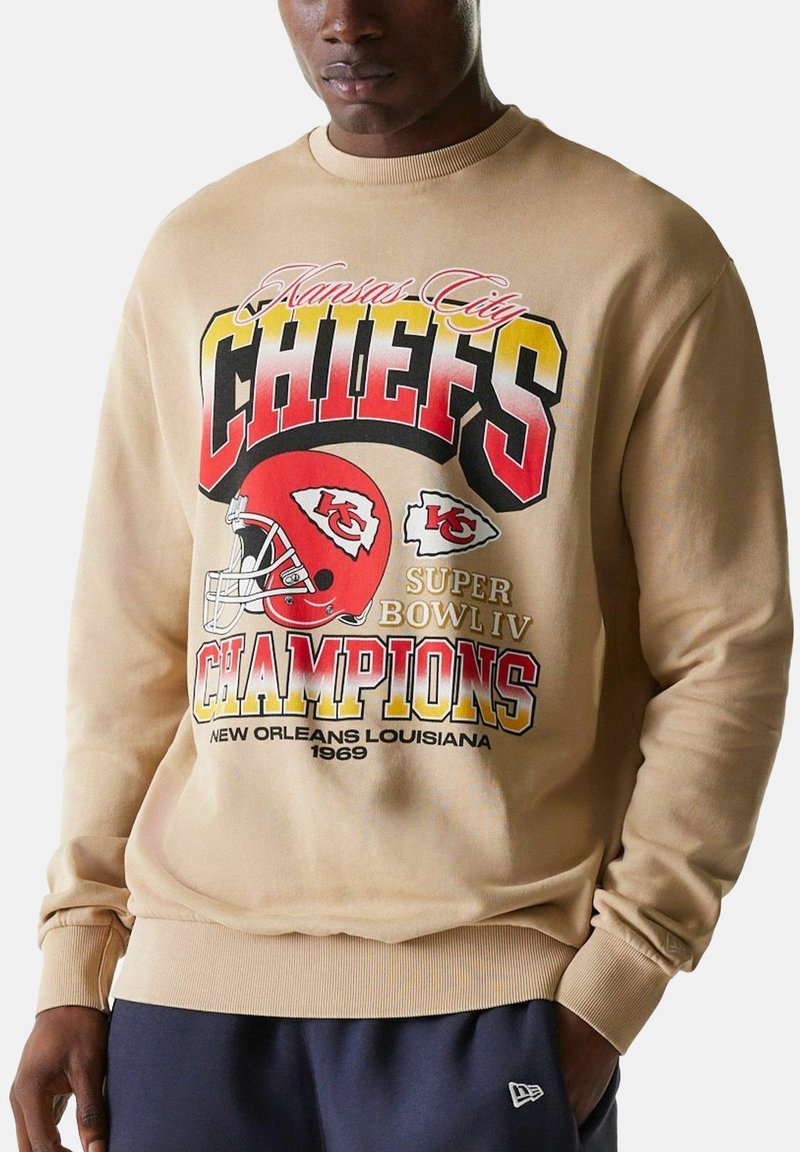 Beige collegepaita, jossa on iso punainen ja keltainen teksti "Kansas City Chiefs Super Bowl IV Champions 1969" ja logo, jota käyttää tummaihoinen henkilö.
