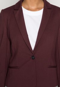 Blazer bordeaux avec revers crantés, fermeture à un bouton noir, et deux poches avant, porté sur un t-shirt blanc.