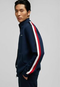 Marinefarbene Zip-Up-Jacke mit rot-, weiß- und blauem Streifen auf den Ärmeln; ausgestattet mit einem hohen Kragen und Fronttaschen; aus weichem, strukturiertem Material.