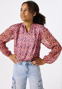 Chemisier coloré à manches longues avec un motif floral dans des teintes de rouge, rose et violet ; se caractérise par un décolleté en V et des poignets élastiques, associé à un jean bleu clair.