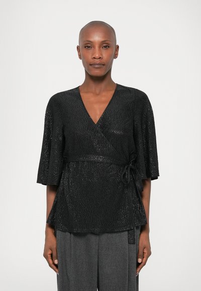 Μαύρο τοπ με υφή, φουσκωτά μανίκια και λεπτομέρεια με ζωνάκι στη μέση, συνδυασμένο με ψηλόμεσες γκρίζες παντελόνες.