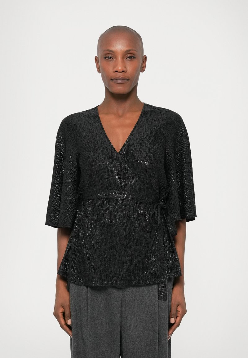 Haut noir à enveloppe avec une surface texturée, des manches évasées et un détail à nouer à la taille, associé à un pantalon gris taille haute.