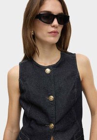 Dunkelblaues Denim-Weste mit Rundhalsausschnitt und goldenen Knopf-Akzenten. Das Model trägt schwarze, kantige Sonnenbrillen, die das strukturierte Design des Kleidungsstücks betonen.