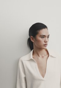 Pull crème, à col en V profond, avec un design à col, doté de longues manches et d'une texture douce, porté par une personne sur un fond clair.