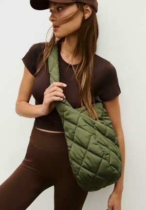 Quilted olive green shoulder bag with a soft texture, featuring a diagonal strap. The model wears a fitted dark brown top and leggings.