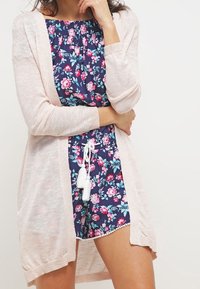 Cardigan lungo rosa chiaro su una tuta con motivo floreale scuro, dotata di cordoncino e dettagli con nappine; vestibilità rilassata e texture morbida.