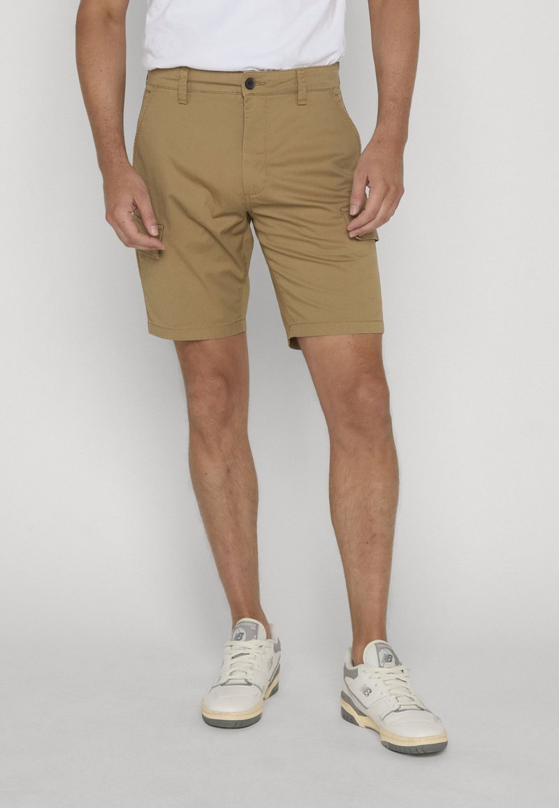 Homme portant un short cargo beige et des baskets blanches, debout devant un fond clair uni.