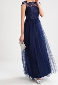 Abito da sera blu navy con corpetto in pizzo, ricami floreali e gonna in tulle. Abbinato a una pochette argentata e scarpe metalliche.