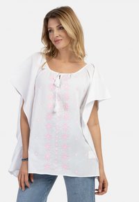 Tunica bianca con ricami floreali rosa, maniche corte e scollatura rotonda. Presenta dettagli decorativi con frange e una vestibilità comoda.