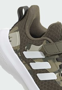 Vihreä kamouflage urheilukenkä, jossa on verkko- ja synteettinen yläosa, kolmea valkoista raitaa ja pehmustettu valkoinen pohja. Mukana on kantapääremmi.