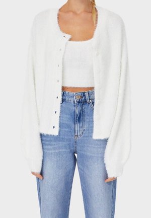 Kvinde iført hvid fluffy cropped cardigan over matchende hvid fluffy croptop og høj talje lyseblå jeans.