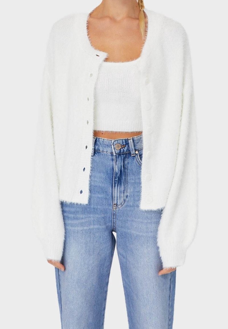 Femme portant un cardigan court duveteux blanc sur un crop top duveteux blanc assorti et un jean taille haute bleu clair.