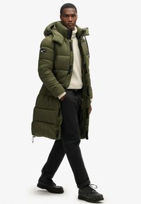 Abrigo acolchado largo verde con capucha, que presenta un diseño acolchado y detalles en negro. Lleva encima de un suéter crema y pantalones negros, con zapatos negros.