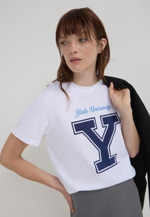 Jeune femme portant un t-shirt blanc de l'université de Yale, tenant une veste noire sur son épaule, regardant sur le côté devant un fond uni.