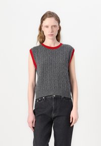 Un pull sans manches en tricot gris avec un motif en torsades et un col et des emmanchures côtelés rouges, associé à un jean noir taille haute.