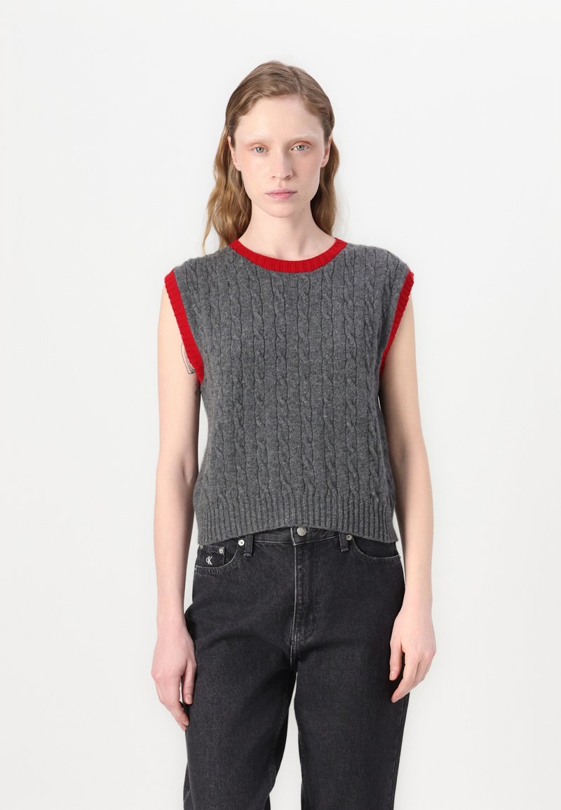 Un pull sans manches en tricot gris avec un motif en torsades et un col et des emmanchures côtelés rouges, associé à un jean noir taille haute.