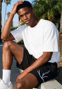 Fiatal férfi fehér pólóban és fekete rövidnadrágban, aki kültéren, egy járdaszegélynél ül, egyik kezét a fején nyugtatva, mögötte pálmafák láthatók.