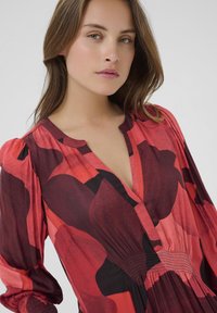 Rotes Blumenkleid mit tiefem V-Ausschnitt, langen Puffärmeln und gerafftem Taillenbereich. Es zeigt große, dunkelrote und schwarze Blumenmuster auf einem hellen Hintergrund.