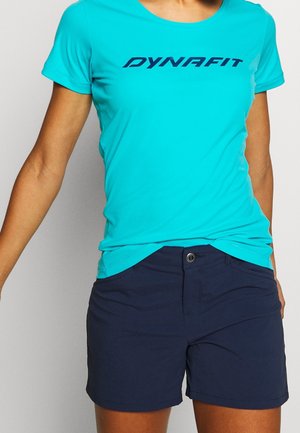 Persona che indossa una maglietta Dynafit a maniche corte di colore azzurro brillante e pantaloncini blu scuro, in piedi davanti a uno sfondo chiaro e uniforme.