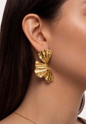 Gros plan sur l'oreille d'une femme portant une grande boucle d'oreille en or texturé ressemblant à deux formes d'éventail superposées.