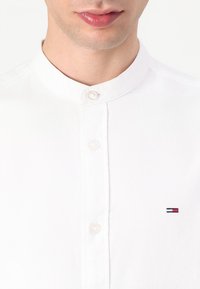 Weißes Baumwollhemd mit Stehkragen, ausgestattet mit einer Knopfleiste und einem kleinen gestickten Logo in Rot, Weiß und Blau auf der linken Seite.