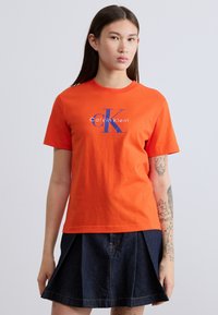 Πορτοκαλί βαμβακερό t-shirt με κοντά μανίκια, στρογγυλή λαιμόκοψη και μπλε τυπωμένο λογότυπο "Calvin Klein" με στιλιζαρισμένο "CK" στο κέντρο.