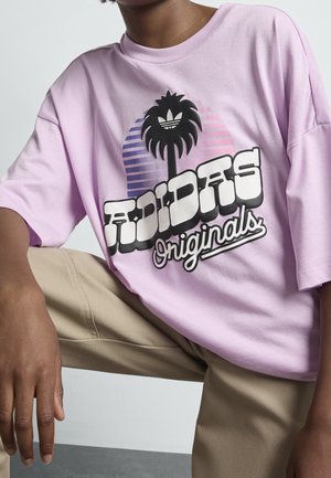 Person iført en lyse lilla Adidas Originals t-shirt med et palmetræsgrafik og blokbogstaver, kombineret med beige bukser.