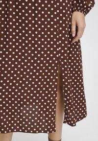 Robe marron à pois blancs, avec une fente latérale et des manches froncées. Le tissu semble lisse avec une texture douce.