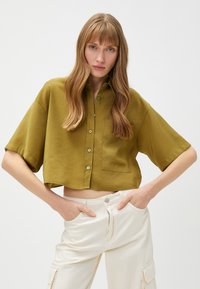 Korte mouwen cropped shirt in olijfgroen stof, met een knoopsluiting aan de voorkant, een enkele borstzak en een gladde textuur.